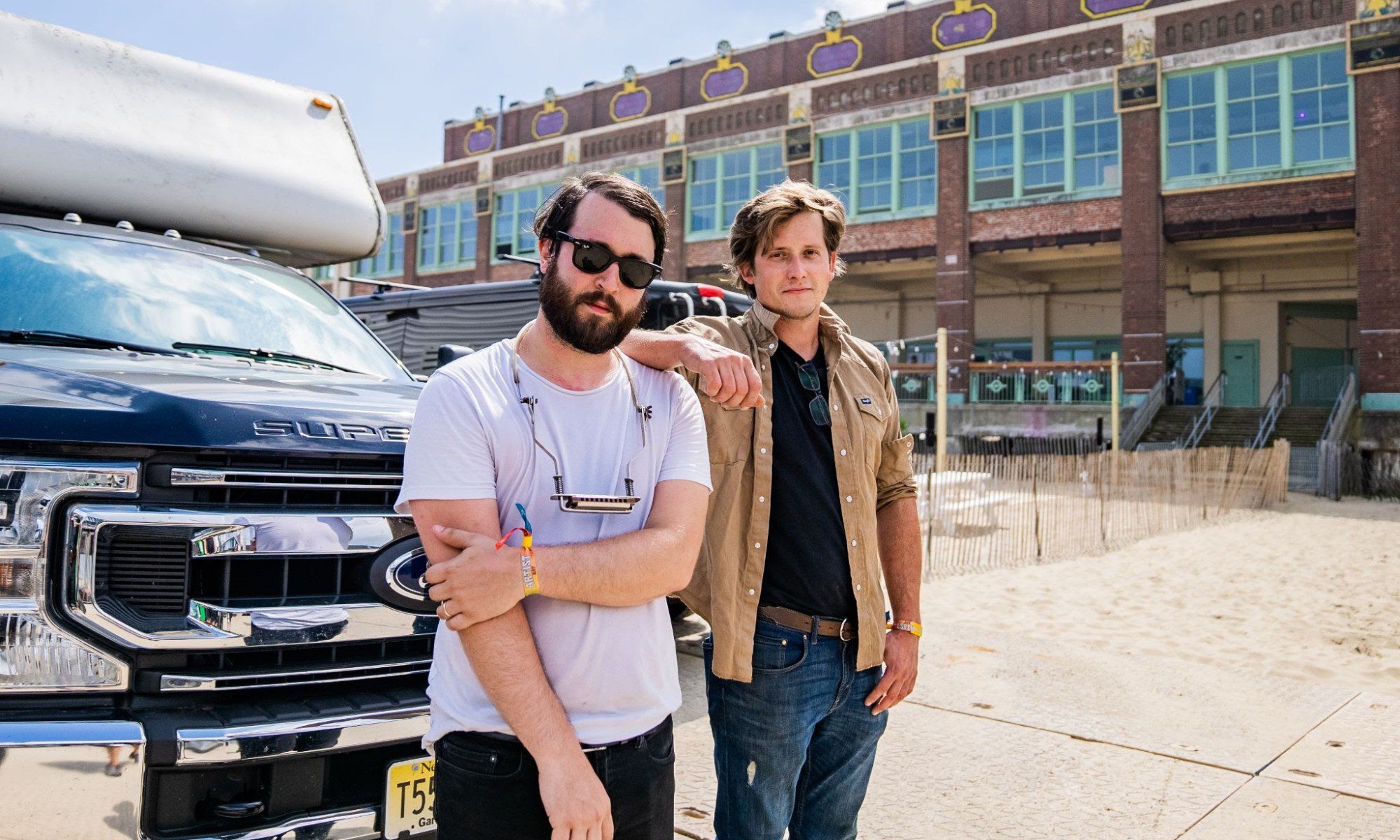 Joe Makoviecki and John Black of the folk band Jackson Pines lean on the front of a pickup. A brick building is in the background.