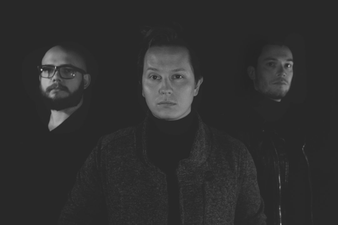 Three men face with minimal expression on their faces in black shirts on a dark background
