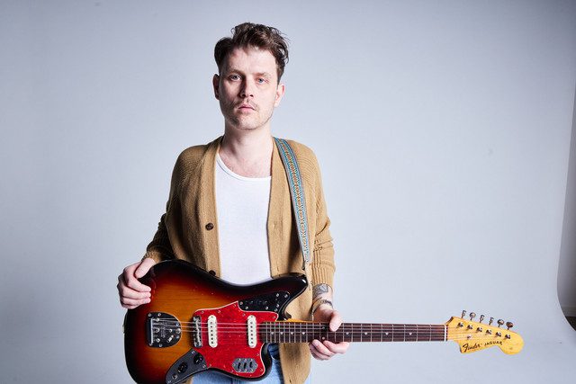 A musician in a cardigan standing with an electric guitar. He is on a neutral background and the look on his face is somber.
